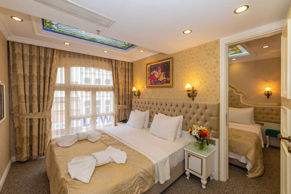 A spacious hotel room with a large bed, a small bedside table with a vase of flowers, and a view of the city through the window.