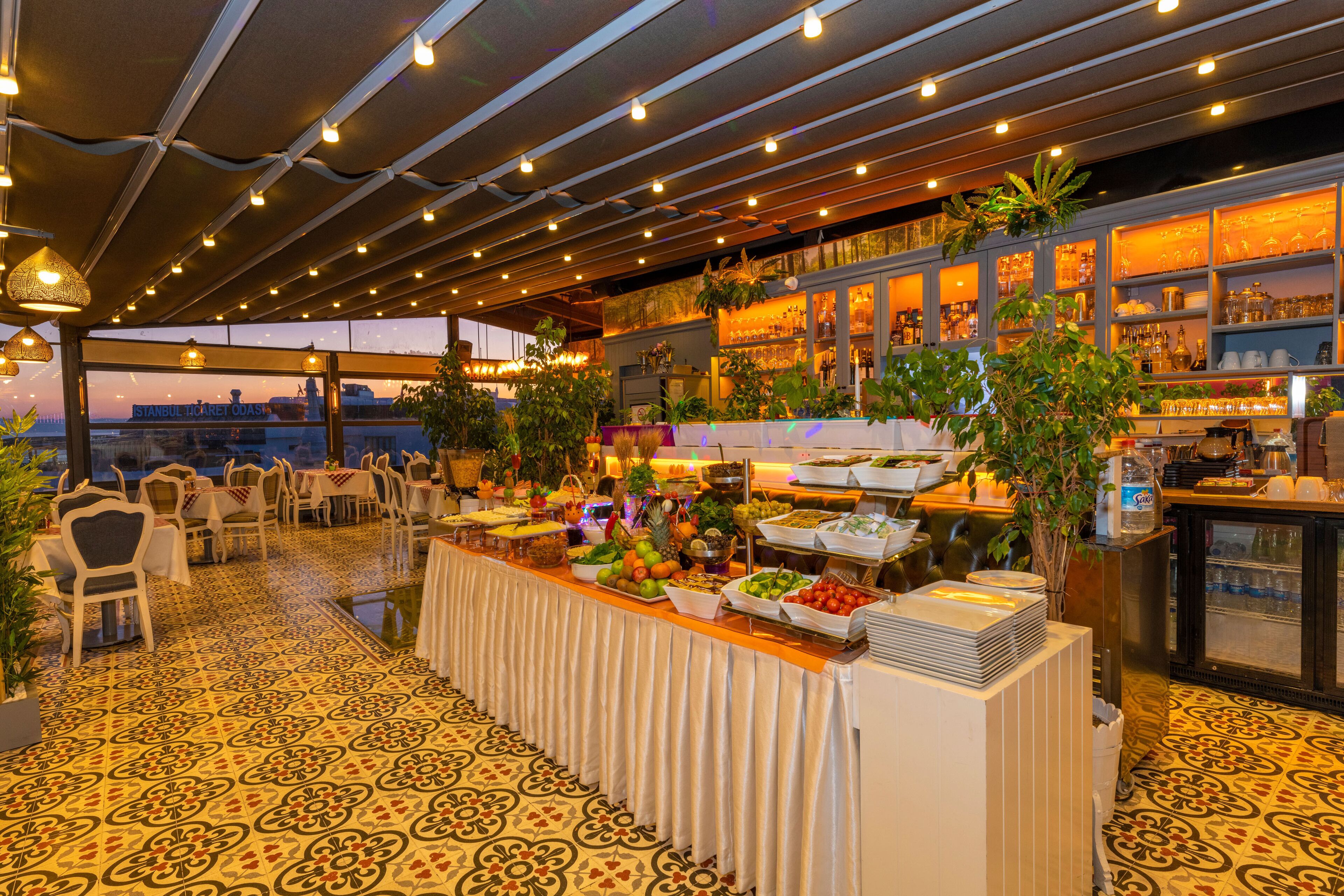 Disposição de buffet com diversos pratos, área de estar com cadeiras e vista para a cidade.