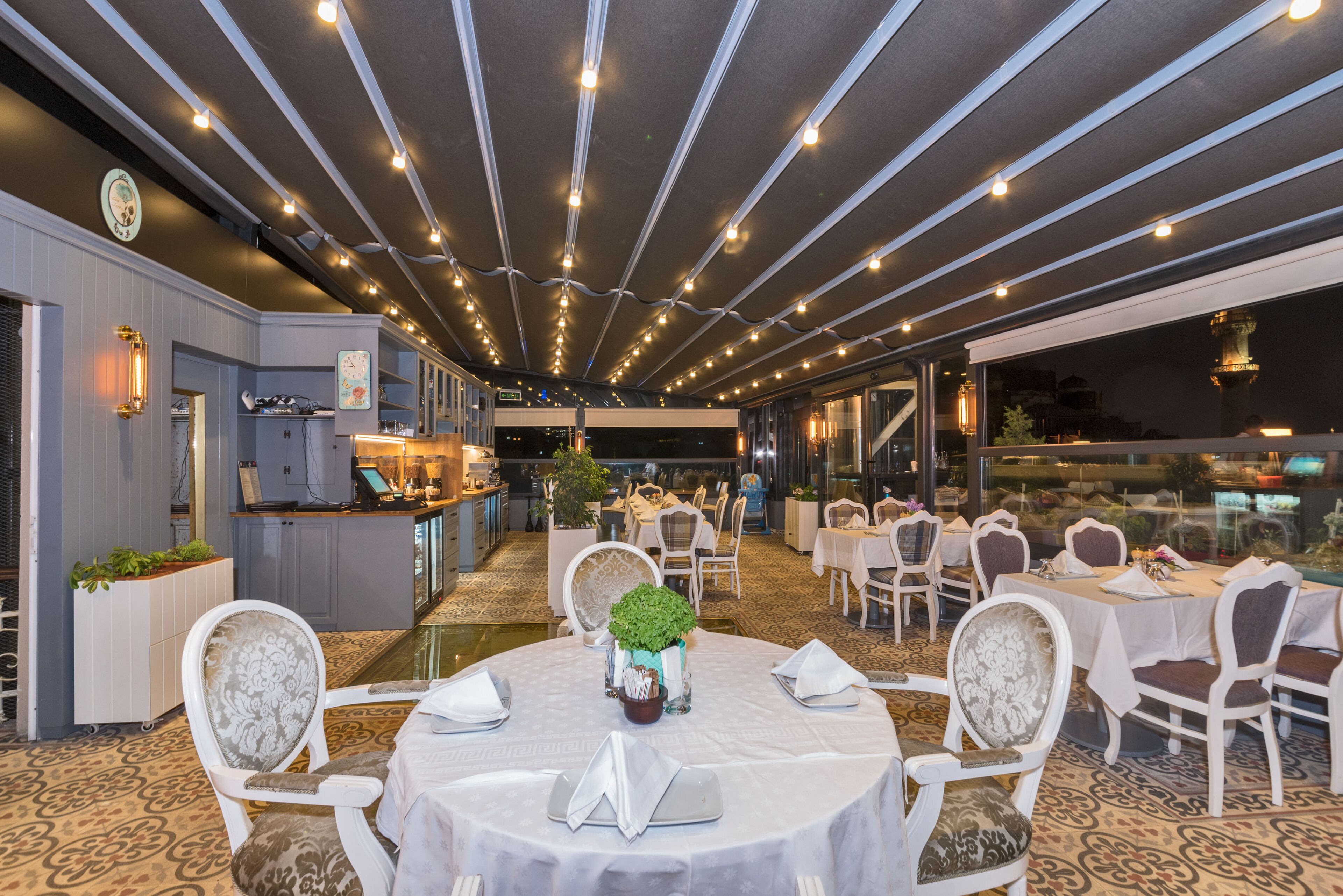 A restaurant with round tables, white chairs, and string lights on the ceiling.