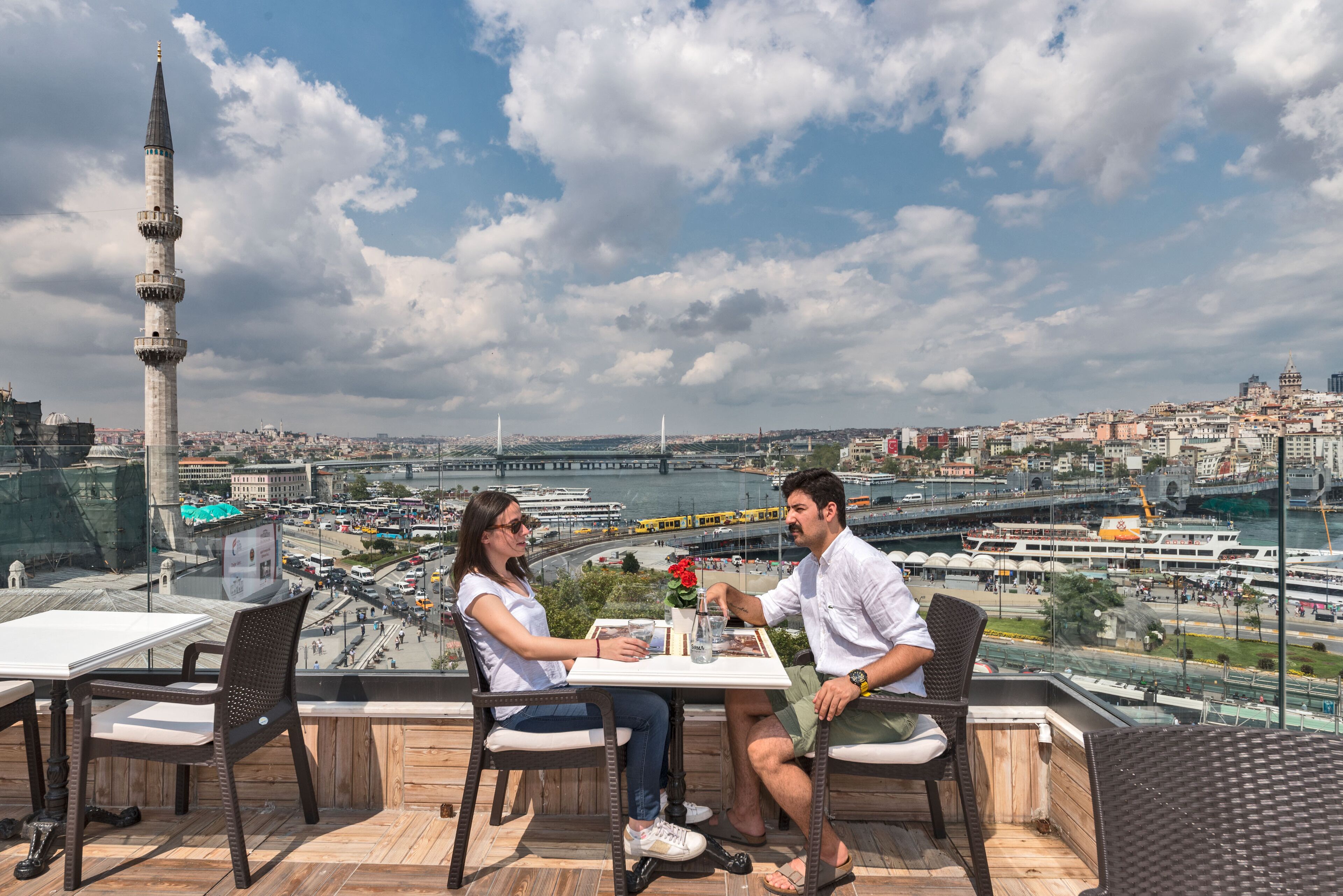 Un couple est assis à une table, dînant et profitant d’une vue sur le paysage urbain, qui comprend une haute tour et un pont.