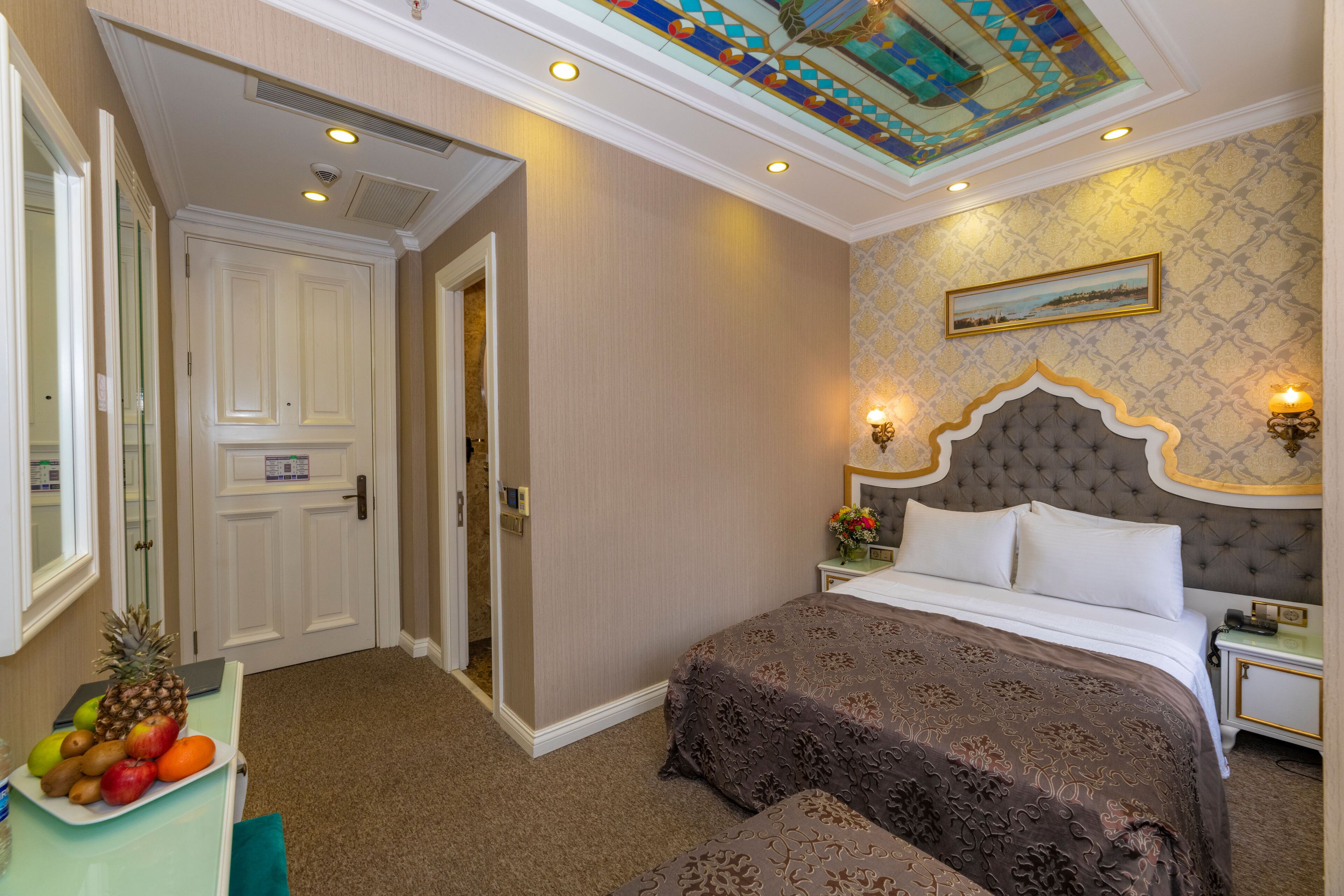 A hotel room with a large bed, a nightstand, a wall-mounted lamp, and a ceiling with a stained glass design.