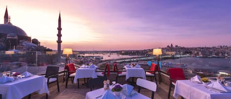 A rooftop restaurant with tables set for dining, overlooking a cityscape and a body of water.