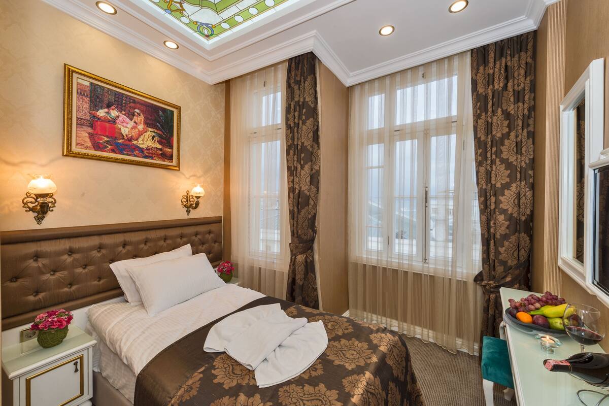 A hotel room with a tufted headboard, a painting above the bed, a nightstand with a vase of flowers, and a table with fruit and a bottle of wine.