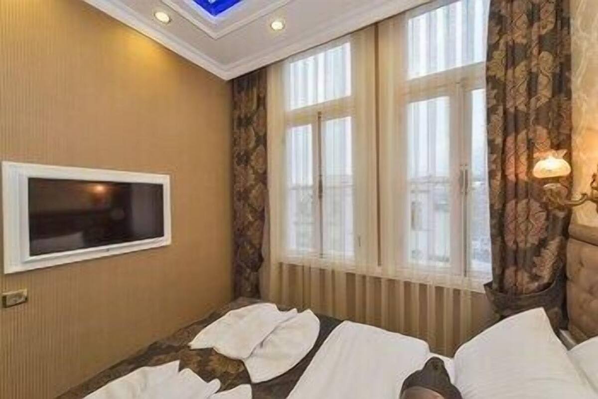 A hotel room with a bed, a flat-screen TV mounted on the wall, a window with curtains, and a ceiling light fixture.