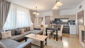 A modern living room with a sofa, dining table, and chairs. There is a kitchen area with a microwave and cabinets. - Fortuna 7 Apartments (Hévíz)