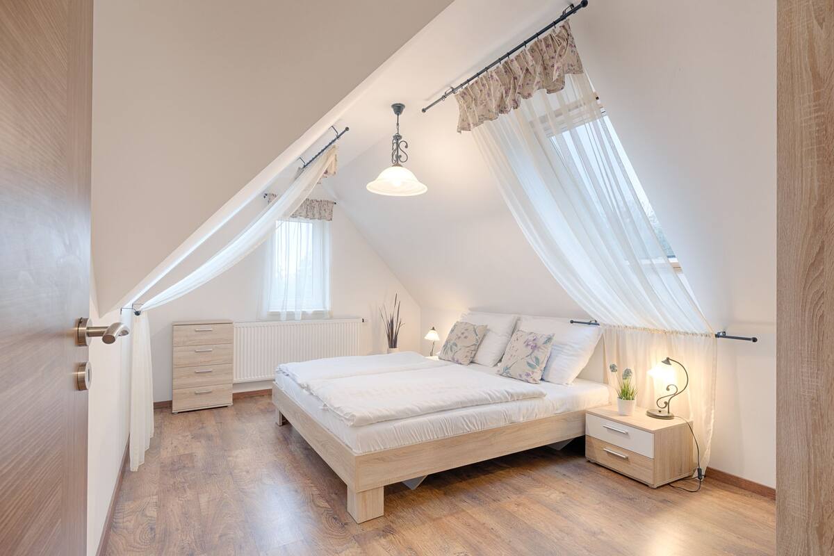 A bedroom with a sloped ceiling, a bed, a bedside table with a lamp, and a radiator.