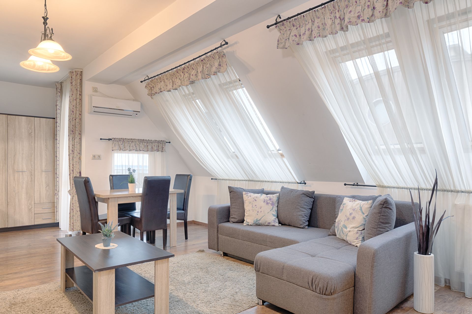 A modern loft apartment with a dining area, sofa, and a large window with sheer curtains.