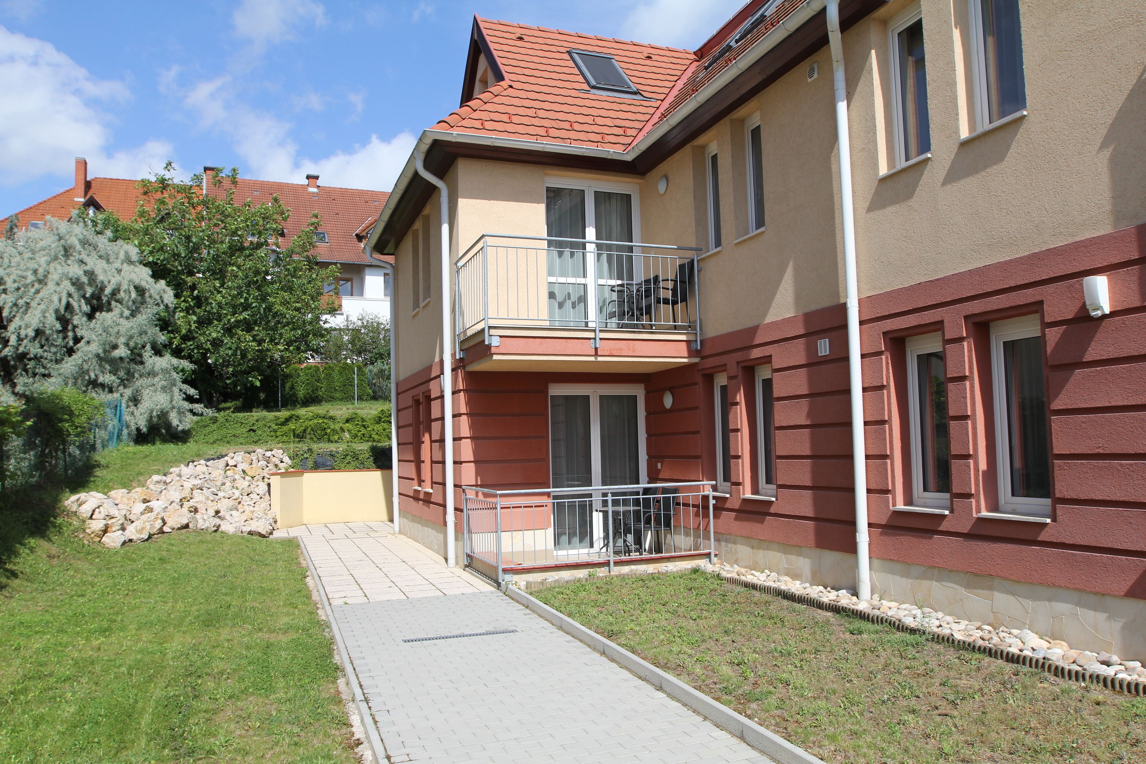 Ein zweistöckiges Wohngebäude mit rotem Ziegeldach, einem Balkon mit Metallgeländer und einer Steinmauer im Vordergrund.