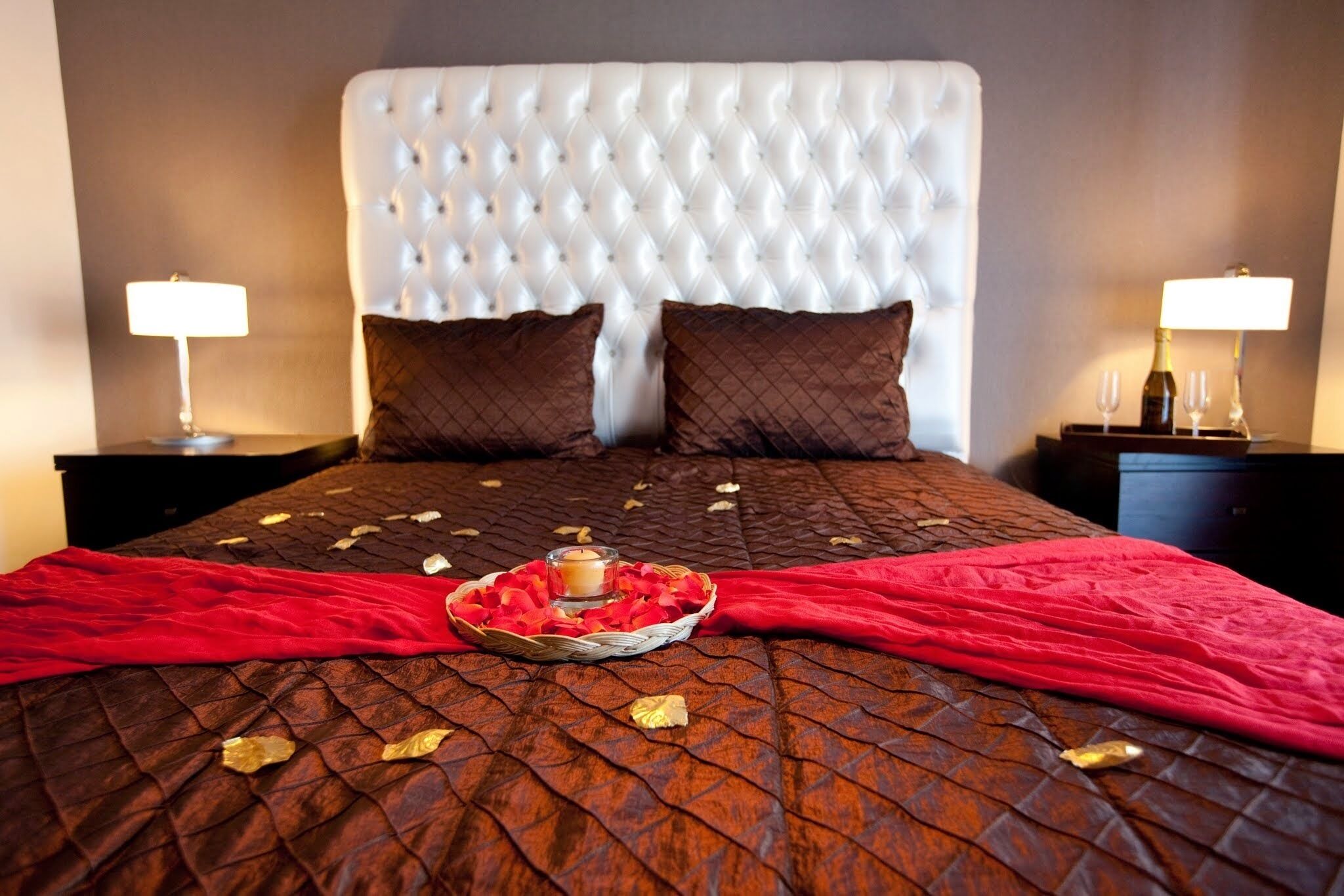 Una cama con cabecera capitonada, dos almohadas, una manta roja, una cesta con pétalos de rosa y una vela, y dos mesitas de noche con lámparas.