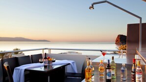 Una terraza con una mesa para dos, un bar con varias botellas, y vistas al mar y a las montañas.