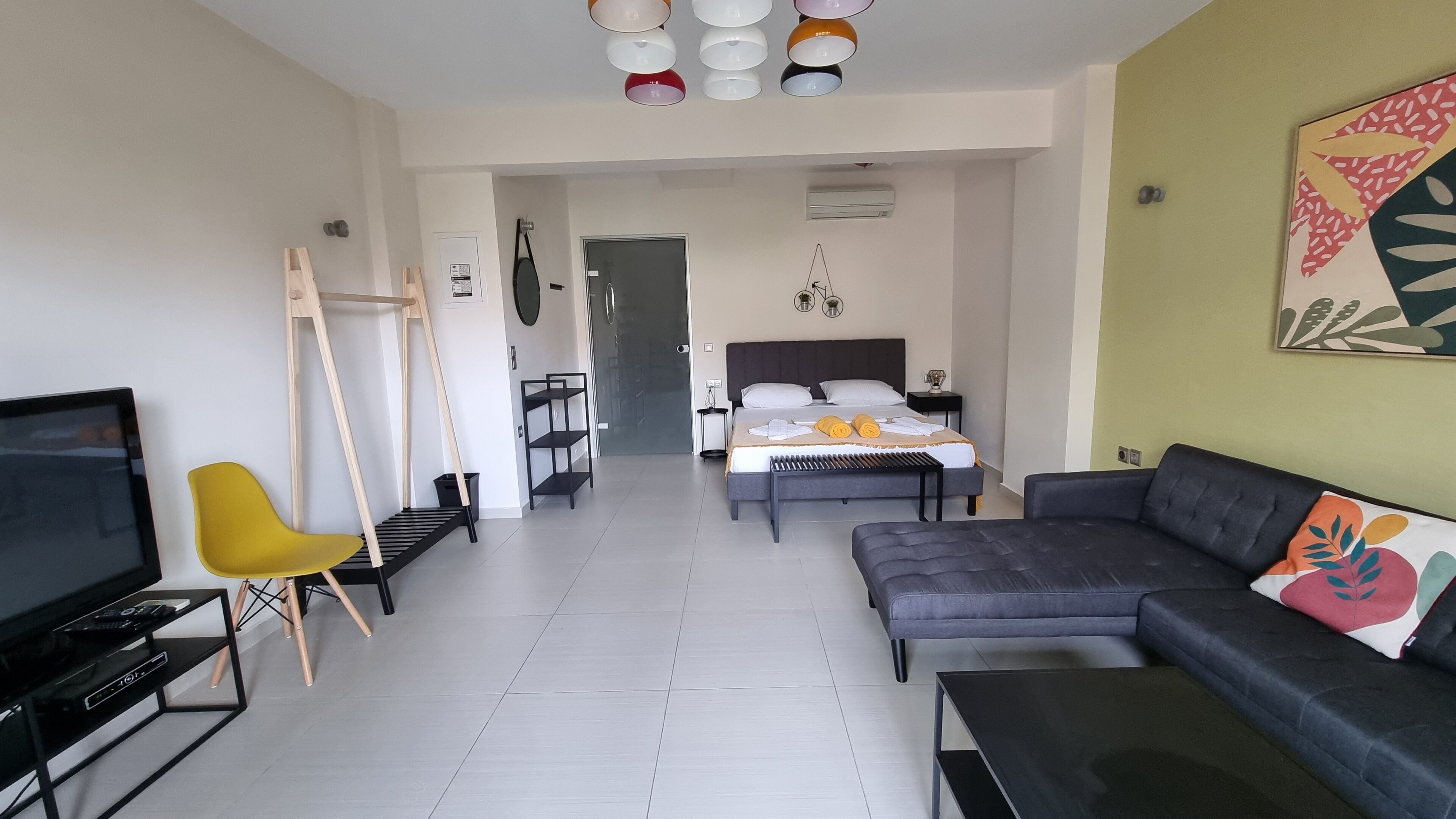 Una sala de estar moderna con un televisor de pantalla plana, un sillón amarillo, un sofá gris y una cama con cabecera blanca.