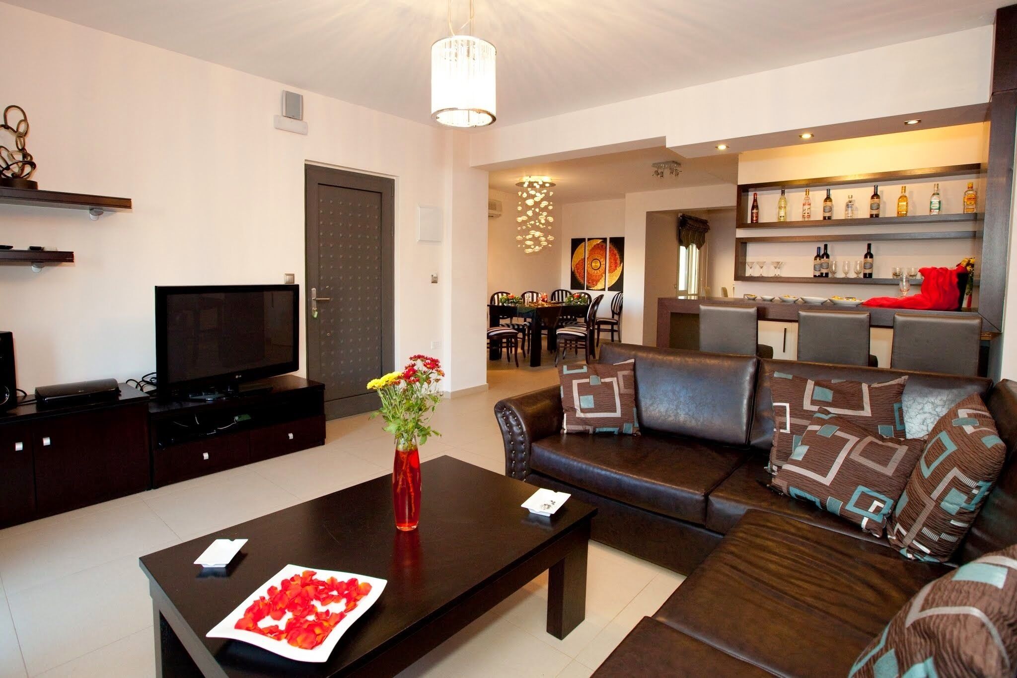 Una sala de estar moderna con un sofá marrón oscuro, una mesa de centro con un florero, un televisor de pantalla plana y una zona de comedor con una araña.