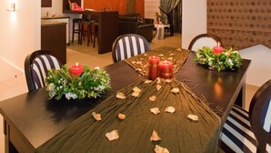 Un comedor con una mesa larga, sillas y una araña. Se ve una cocina al fondo.