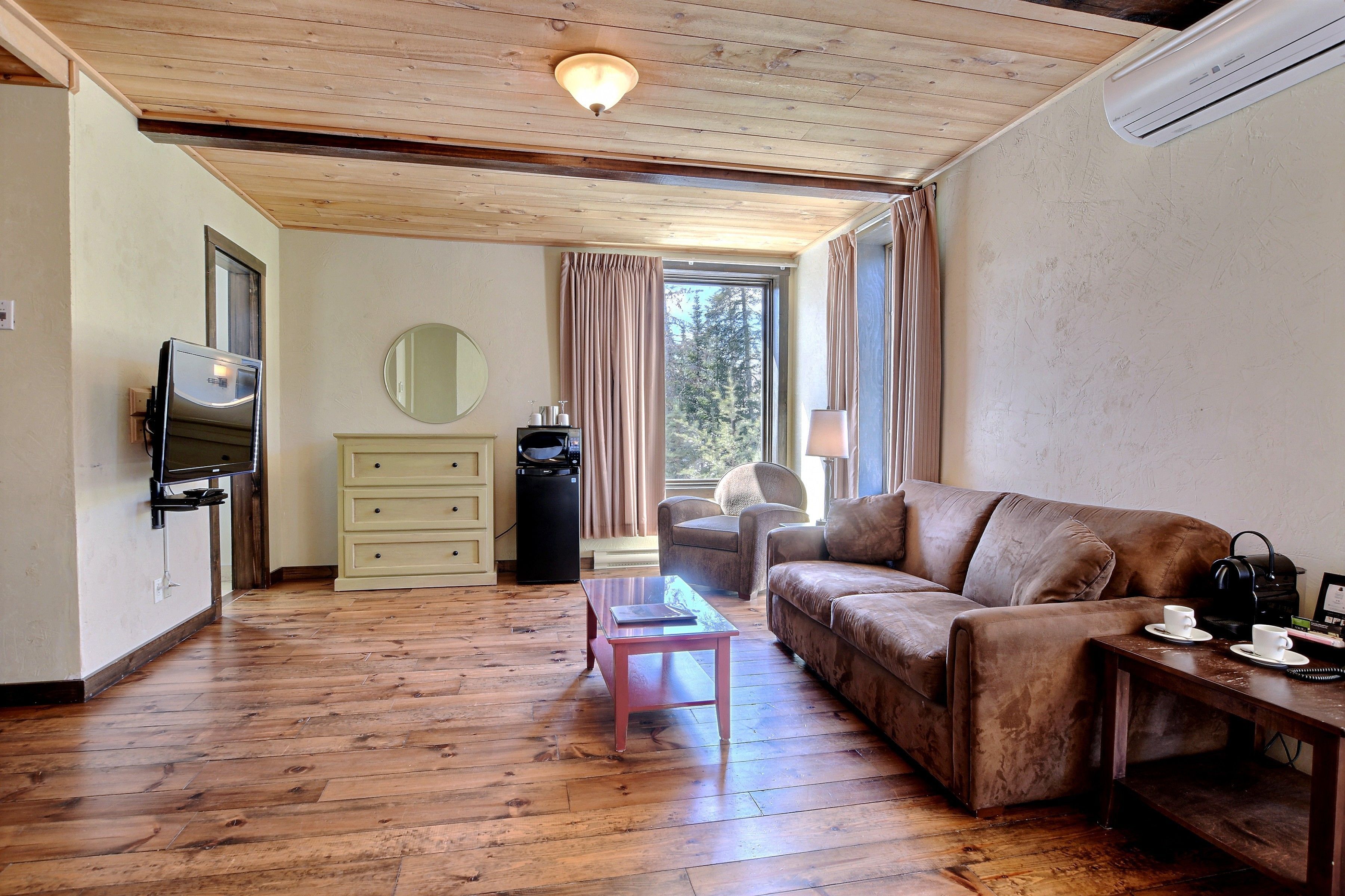 A room with a wooden floor, a brown sofa, a small table, a TV, a mirror, and a window with curtains.
