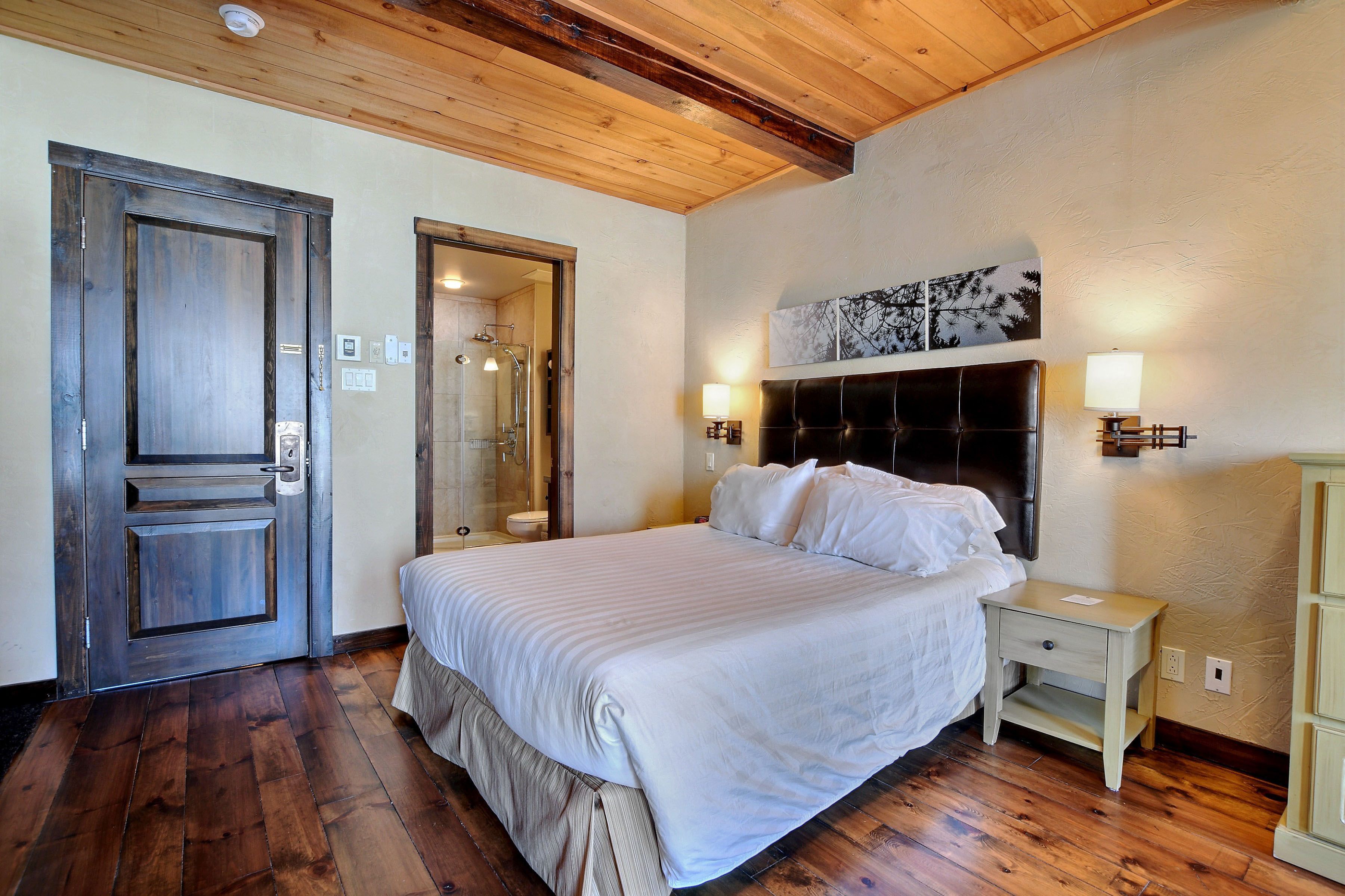Un dormitorio con una cama grande, piso de madera, techo de madera y una mesita de noche con lámpara.