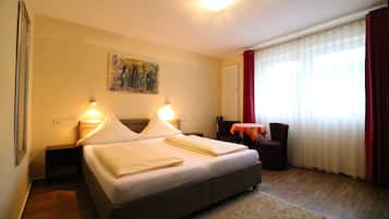 Ein Hotelzimmer mit Bett, Nachttischlampen, einem Bild an der Wand, einem Fenster mit Vorhängen und einem kleinen Tisch mit Stuhl.