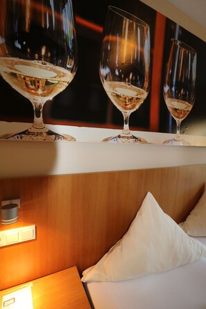 A hotel room with a wooden headboard, a white bed, and three wine glasses on a shelf. - Christiana's Wein & Art Hotel (Bernkastel-Kues)