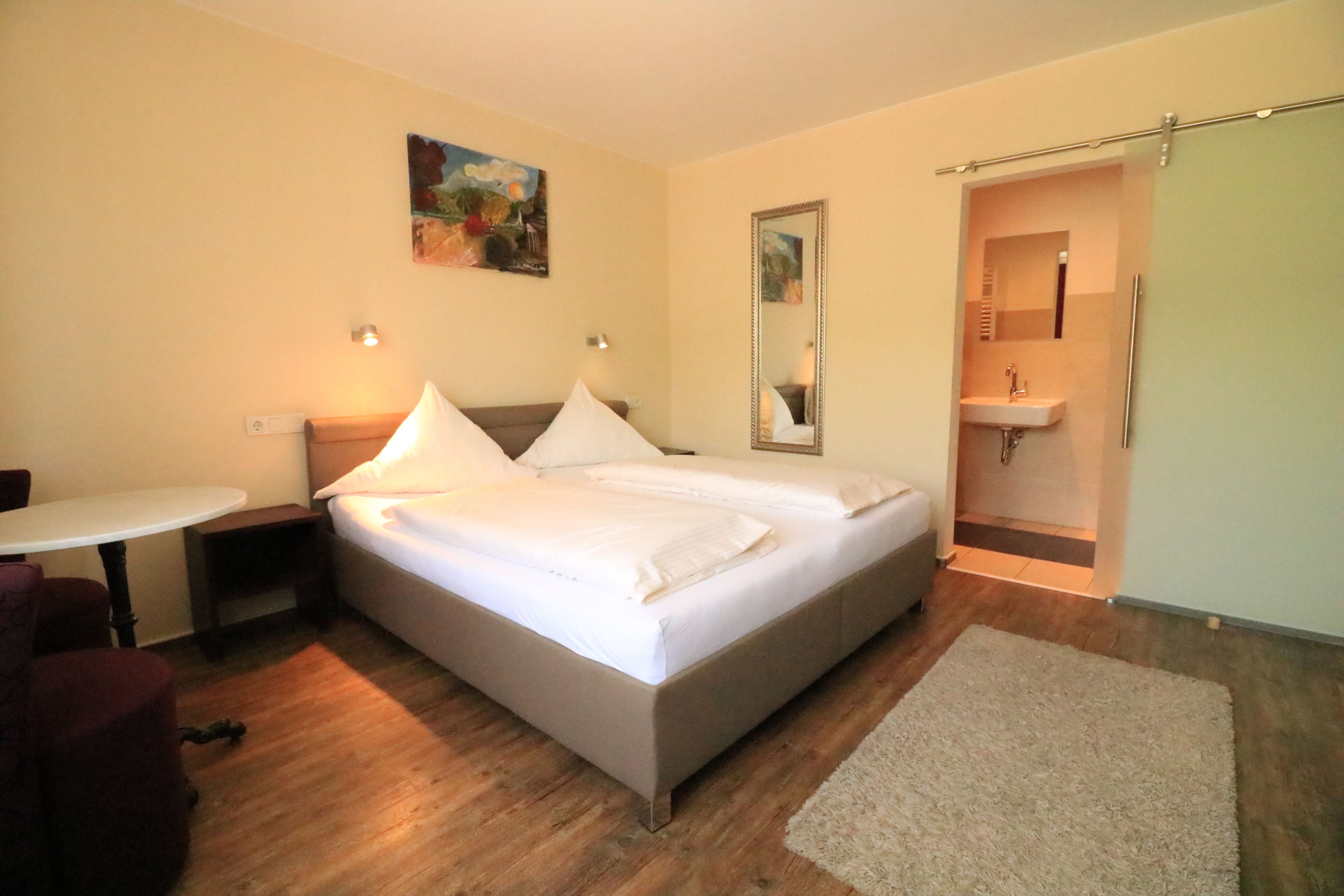 A bedroom with a double bed, bedside tables, a small table, a chair, a mirror, and a bathroom with a sink and shower.