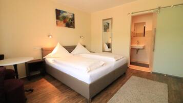 Une chambre à coucher avec un lit double, des tables de chevet, une petite table, une chaise, un miroir et une salle de bain avec un lavabo et une douche.