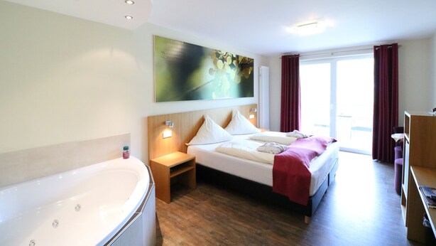 Habitación de hotel con una cama grande, zona de estar, bañera y un ventanal con cortinas.