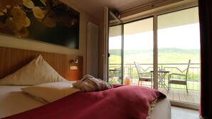 Un cuarto de hotel con una cama grande, una colcha roja, un cuadro en la pared y un balcón con vistas al campo.