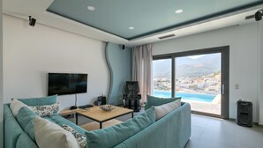 A modern living room with a teal sofa, a flat-screen TV, a coffee table, and a view of a pool and mountains.