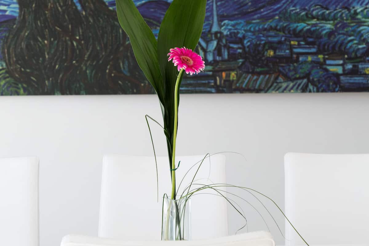 A room with a painting of a night sky and a vase with a pink flower on a white table.