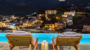 A poolside area with wooden lounge chairs and a night light, overlooking a hillside town with illuminated buildings.