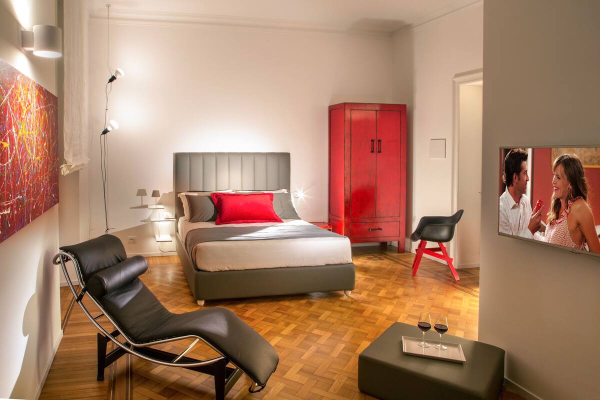 A modern bedroom with a bed, a red wardrobe, a black lounge chair, and a small table with glasses.