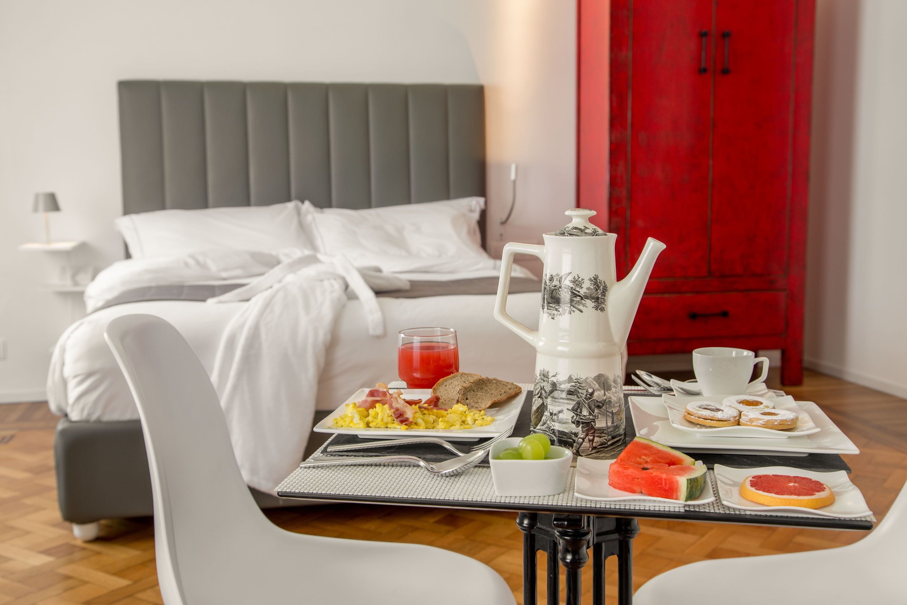 A neatly made breakfast table with a variety of food items, a bed with white linens, and a red cabinet in the background.