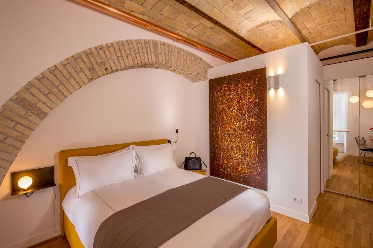 A modern hotel room with a brick archway, a large bed, a wall art piece, and a wooden floor.