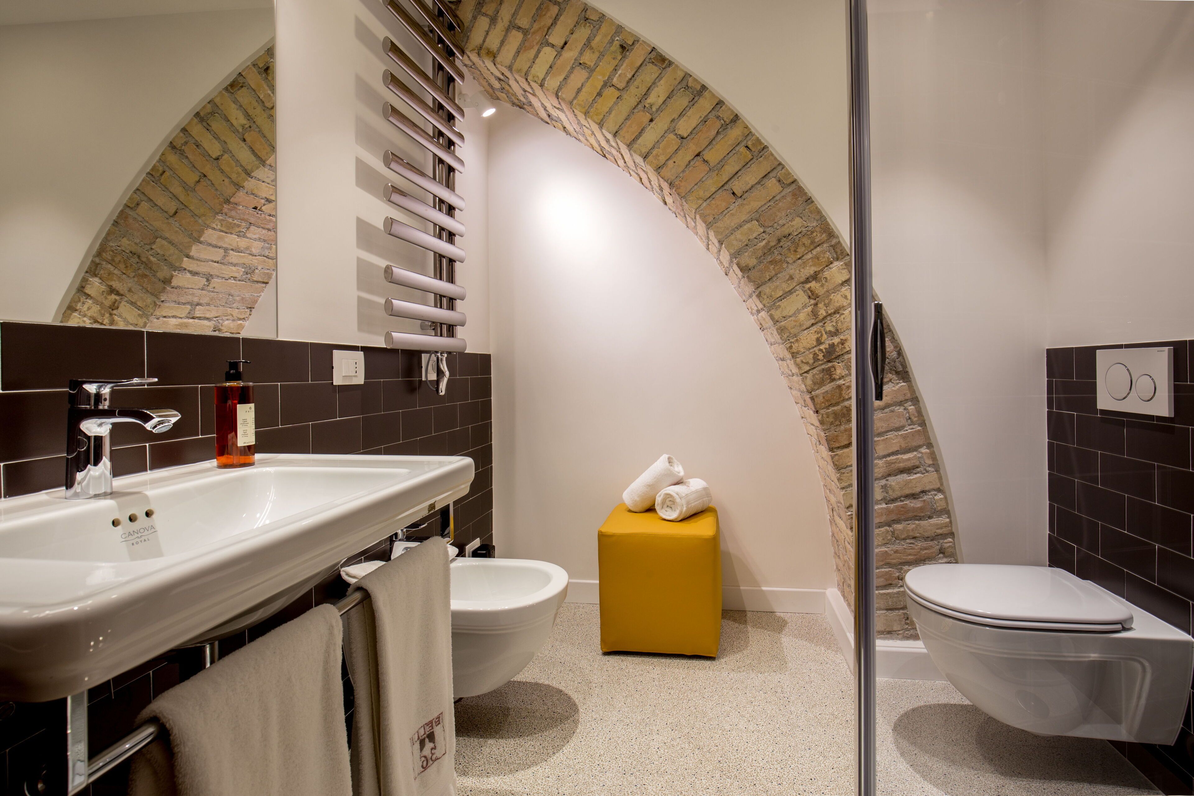 A modern bathroom with a large sink, two toilets, a yellow stool, and a brick archway.