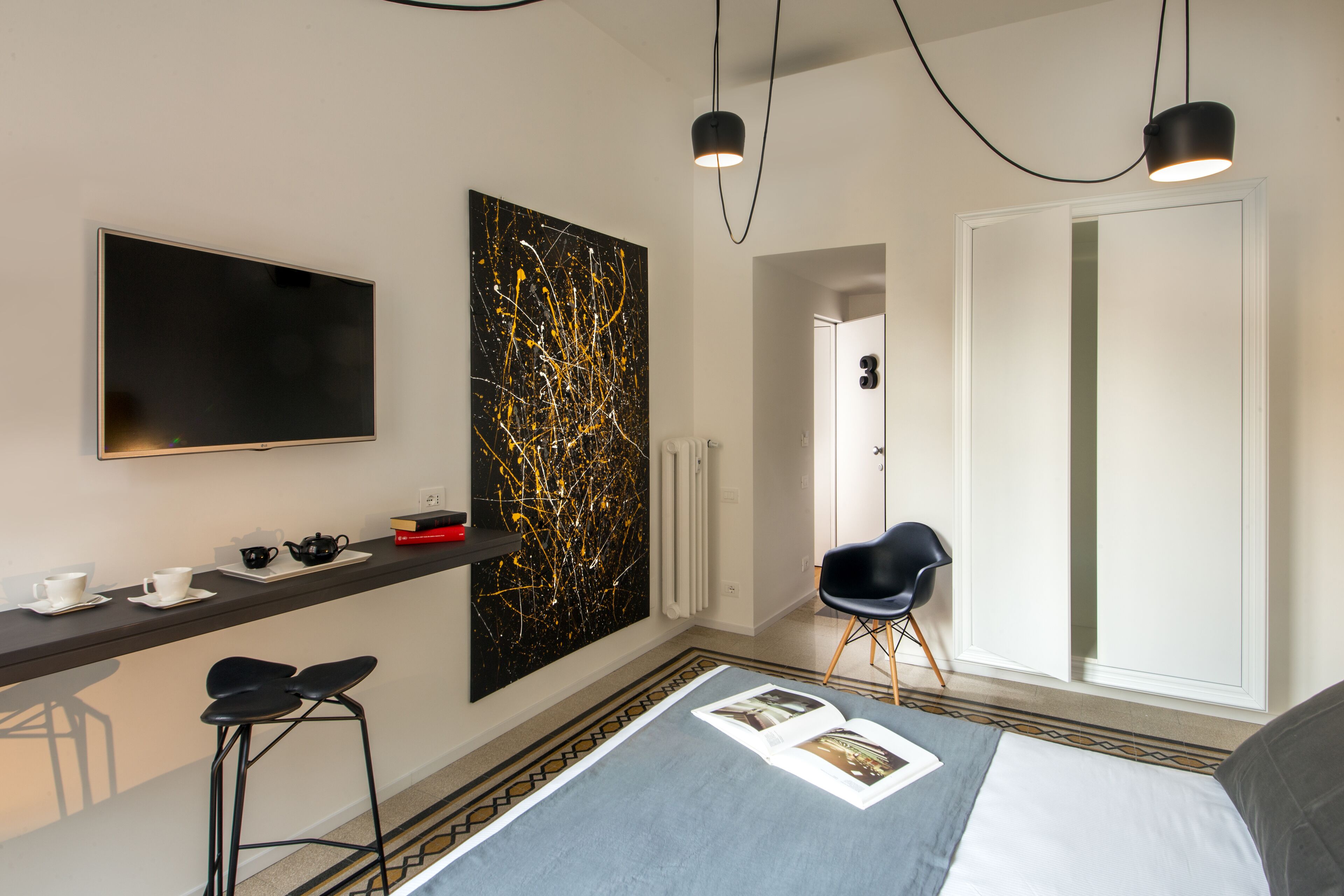 Et moderne hotelværelse med et fladskærms-tv, et stort abstrakt maleri, et barområde med barstole, en seng med et magasin, og en dør der fører til et andet værelse.