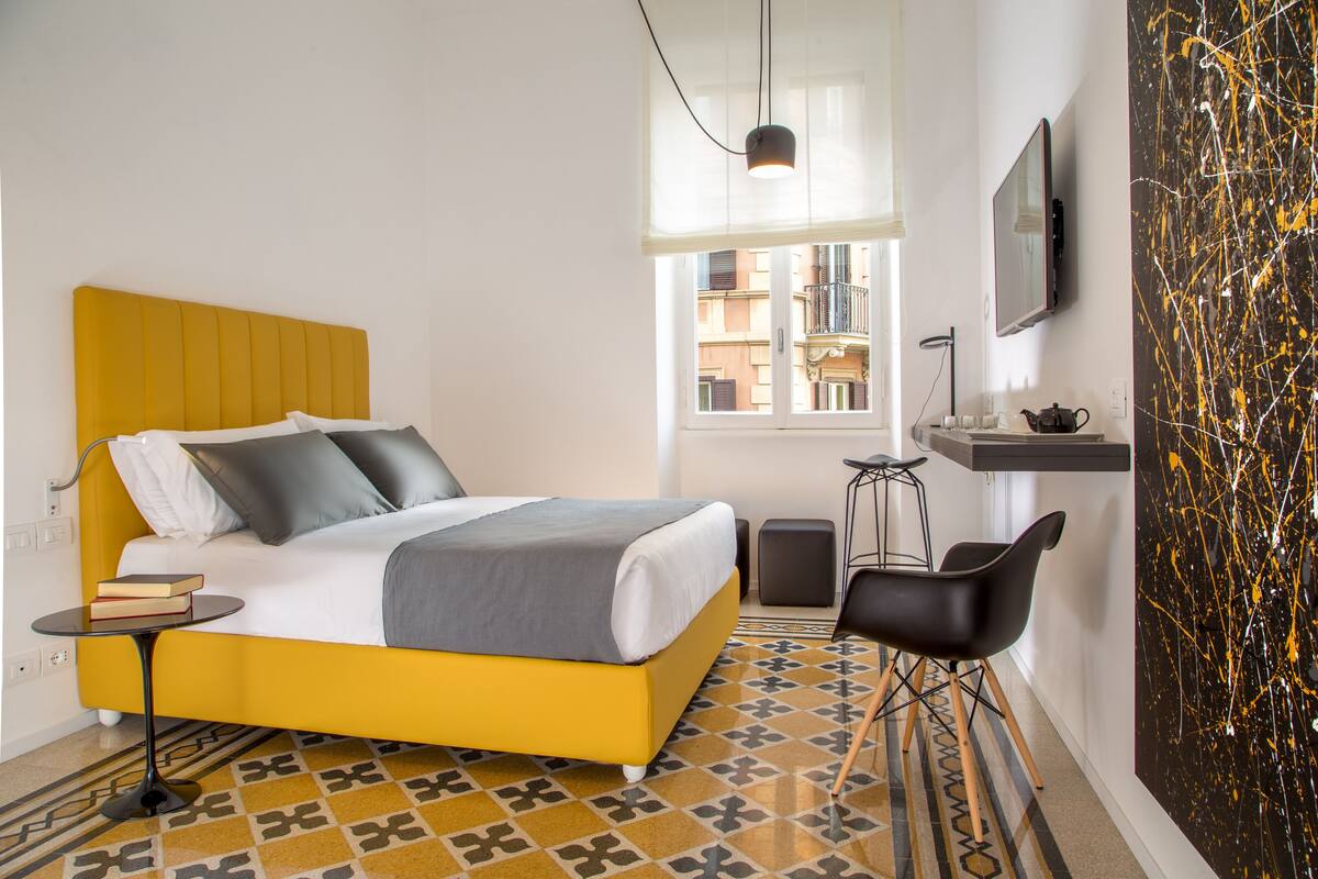 A modern hotel room with a yellow bed, a small desk with a chair, a large window, and a black and white abstract painting on the wall.