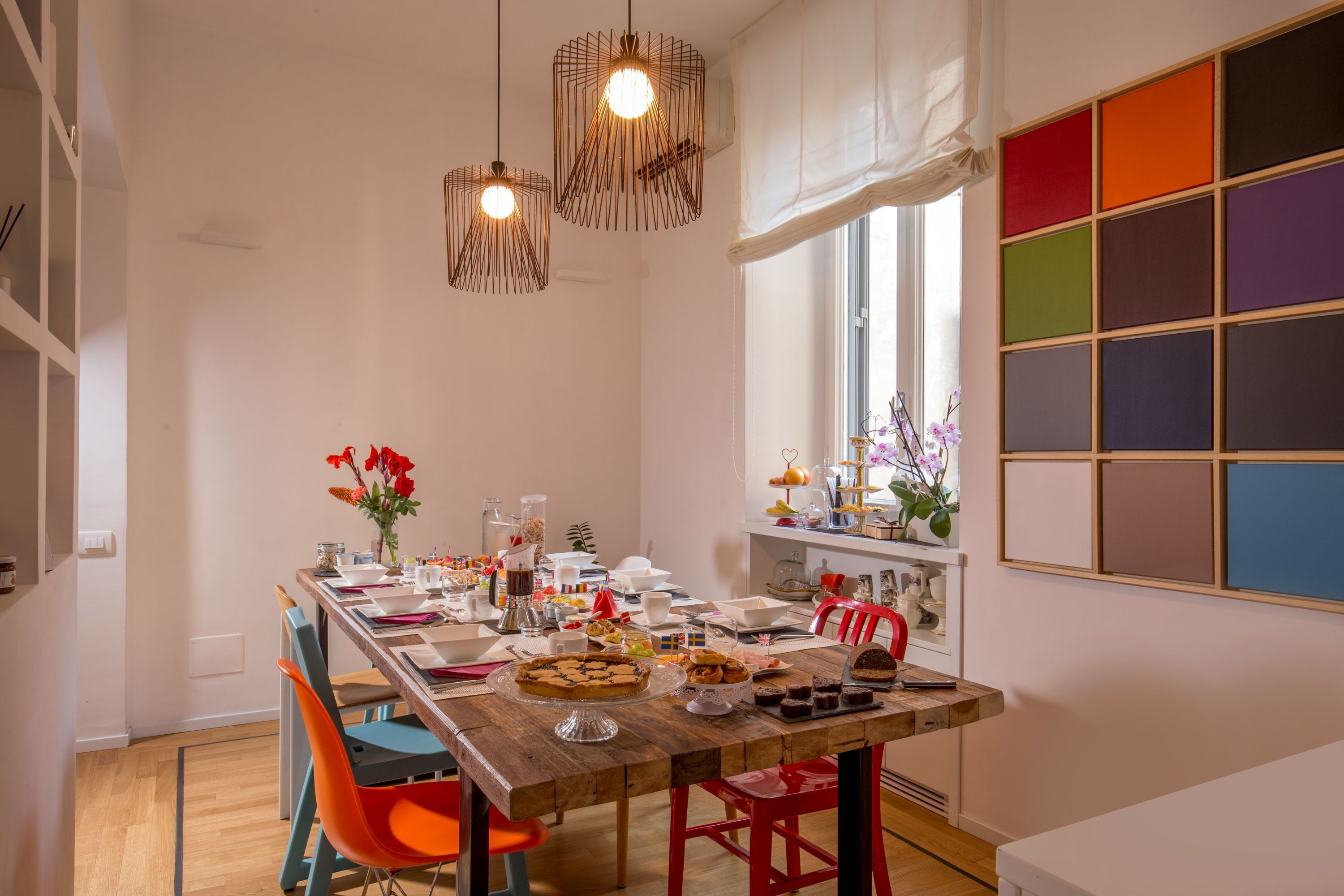 A dining area with a wooden table set for a meal, surrounded by colorful chairs, and a large window with a view.