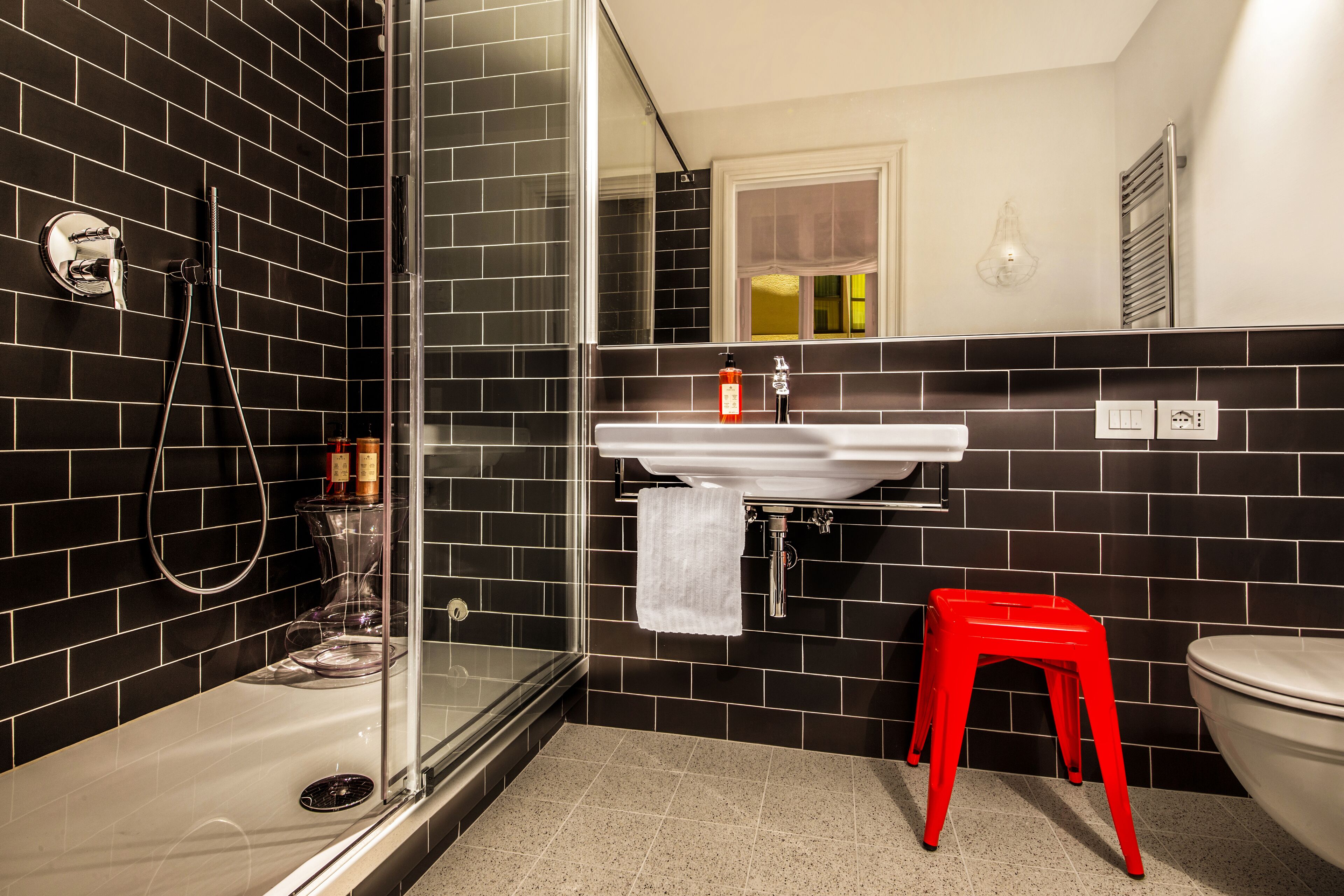 Ein modernes Badezimmer mit schwarzem Wandfliesen, einer Duschkabine aus Glas, einem weißen Waschbecken und einem roten Hocker.