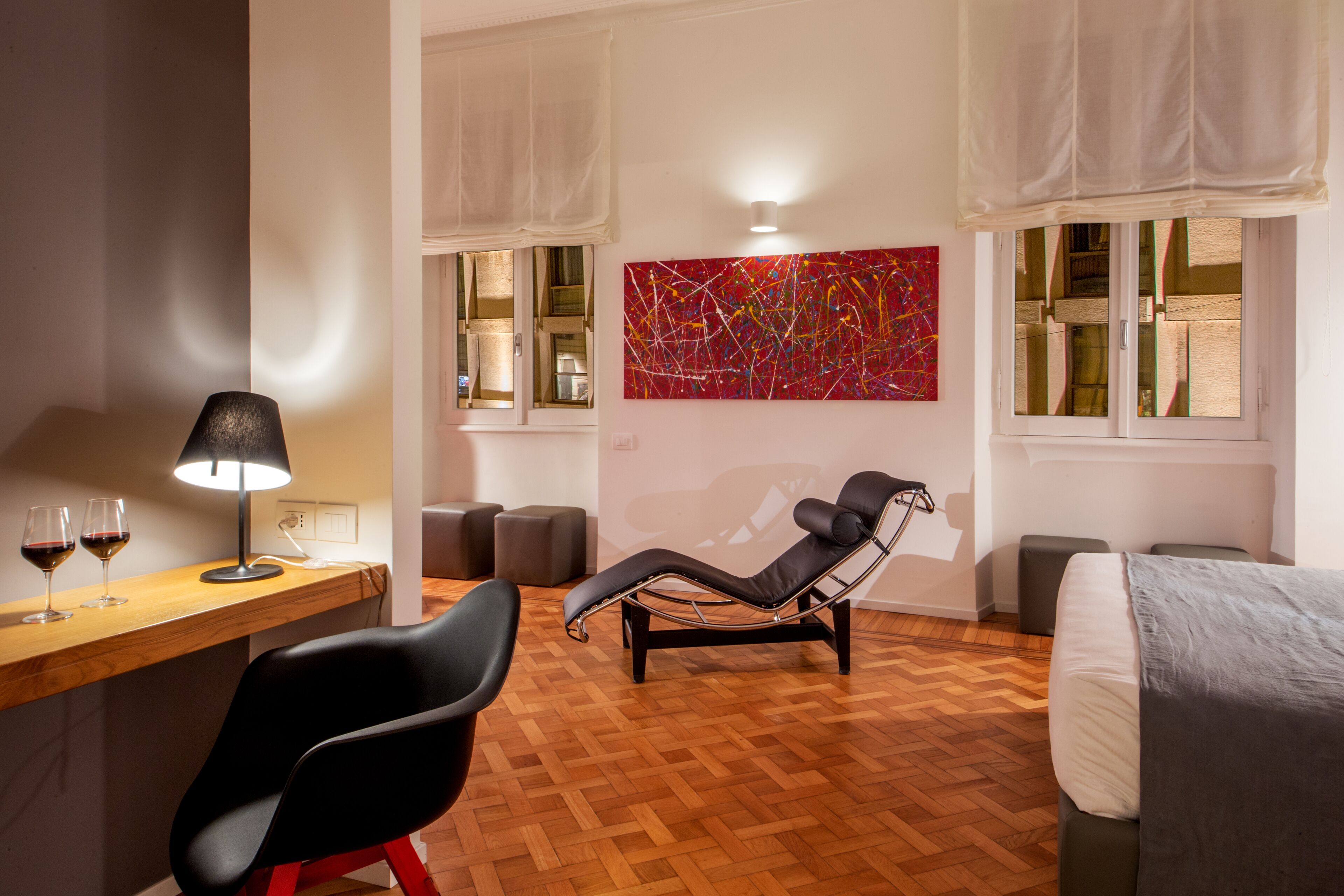 A modern hotel room with a wooden floor, a black lounge chair, a desk with a lamp, and a large abstract painting on the wall.