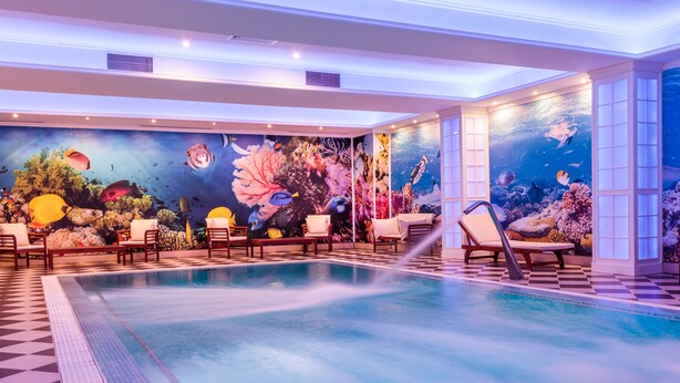 Una amplia piscina interior con suelo a cuadros, rodeada de sillas de descanso y un mural con temática marina.