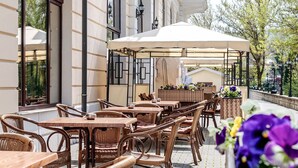 Un café al aire libre con mobiliario de mimbre y sombrillas, frente a un edificio con ventanas amplias y una exhibición de flores.