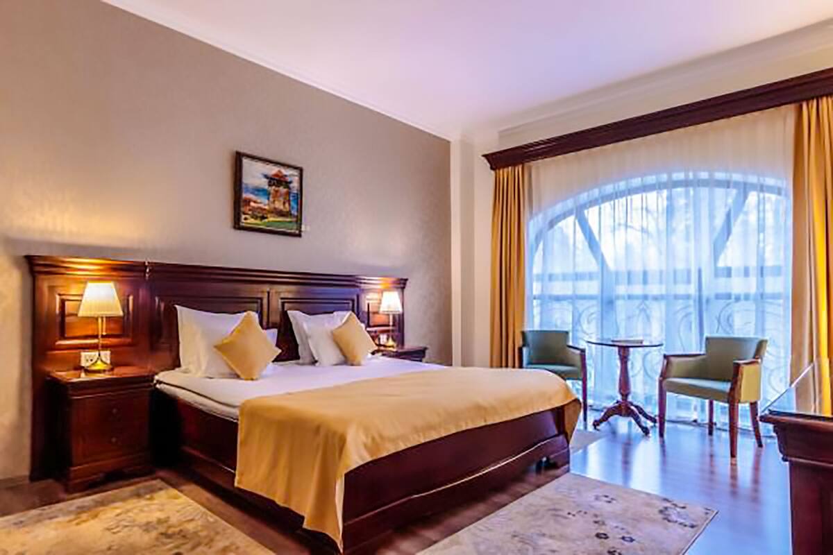 A hotel room with a large bed, two bedside tables with lamps, a small table and chairs, and a large window with curtains.