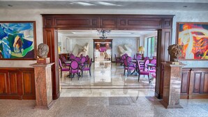 Un vestíbulo amplio con suelo de mármol, sillas moradas y una mesa central. Se observan grandes cuadros en las paredes y un restaurante al fondo.