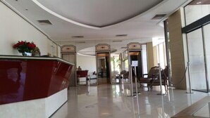 Zona de recepción de un hotel con un mostrador rojo, una pared de vidrio y un área de descanso con sillas y una mesa.
