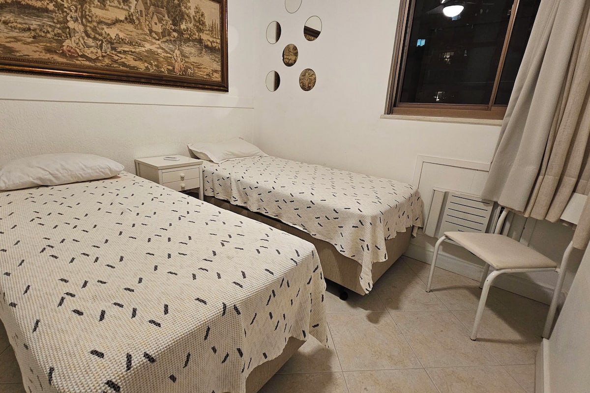 Un dormitori amb dos llits, una taula de nit, una cadira, un radiador i una finestra amb cortines.