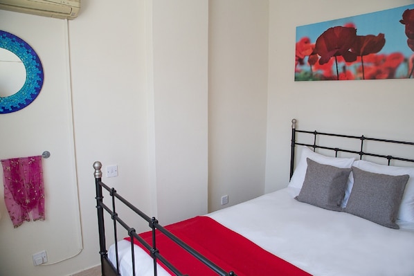 A neatly made bed with a red blanket and two pillows, a round mirror with a blue frame, and a painting of red flowers on the wall.