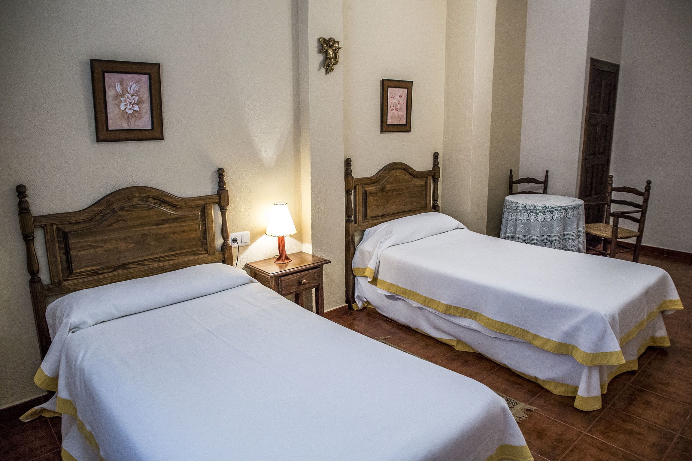 Two single beds with wooden headboards, a small bedside table with a lamp, and a chair at a small round table.