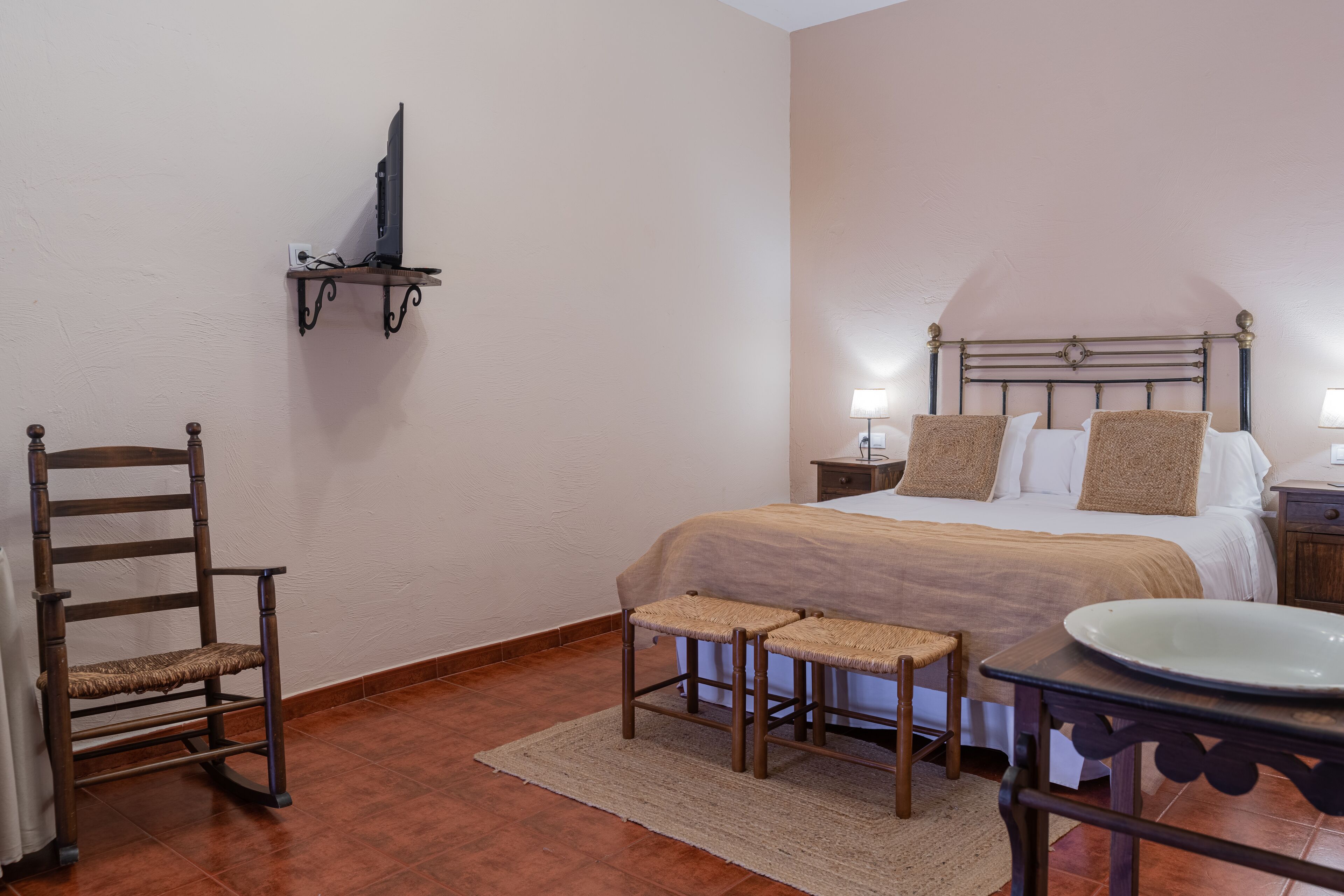 Un dormitorio con cama, mesitas de noche, una silla, un taburete pequeño, un televisor sobre una estantería y un plato decorativo sobre una mesa.