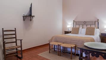 Un dormitorio con cama, mesitas de noche, una silla, un taburete pequeño, un televisor sobre una estantería y un plato decorativo sobre una mesa.