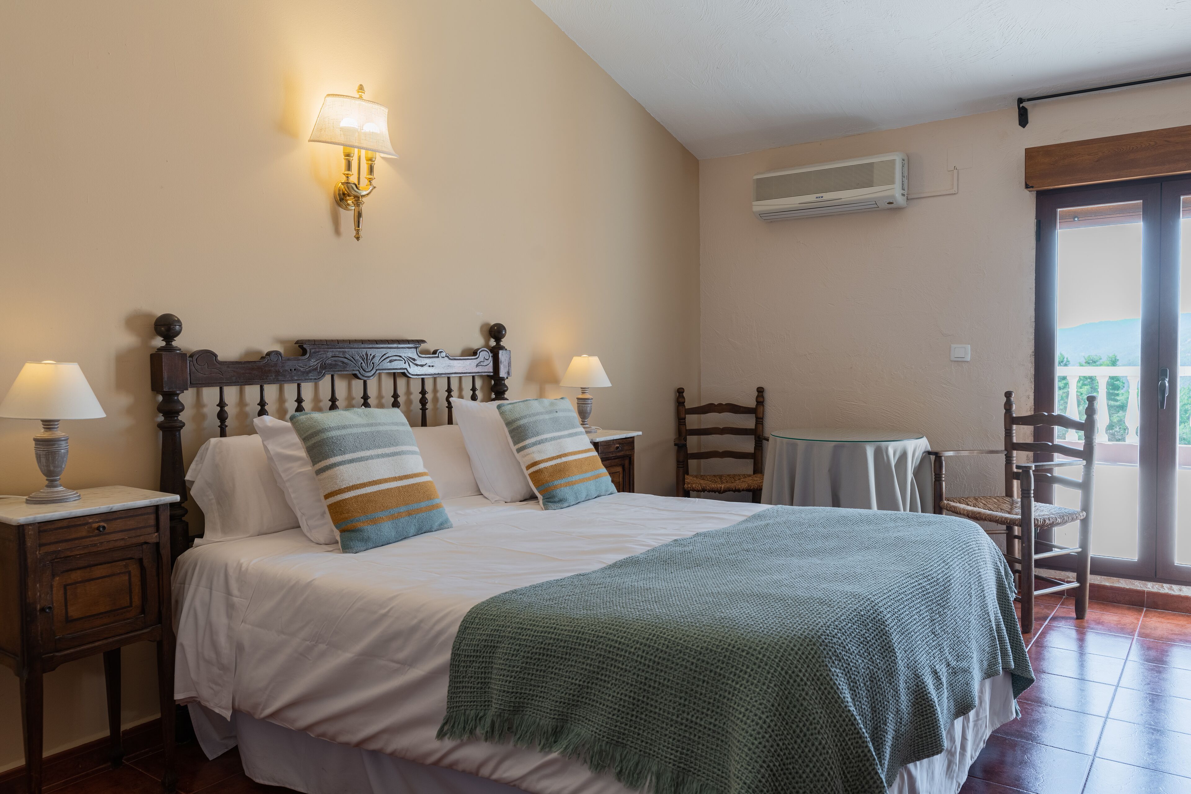 Un dormitorio con una cama, dos mesitas de noche con lámparas, una silla, una pequeña mesa y un equipo de aire acondicionado en la pared.