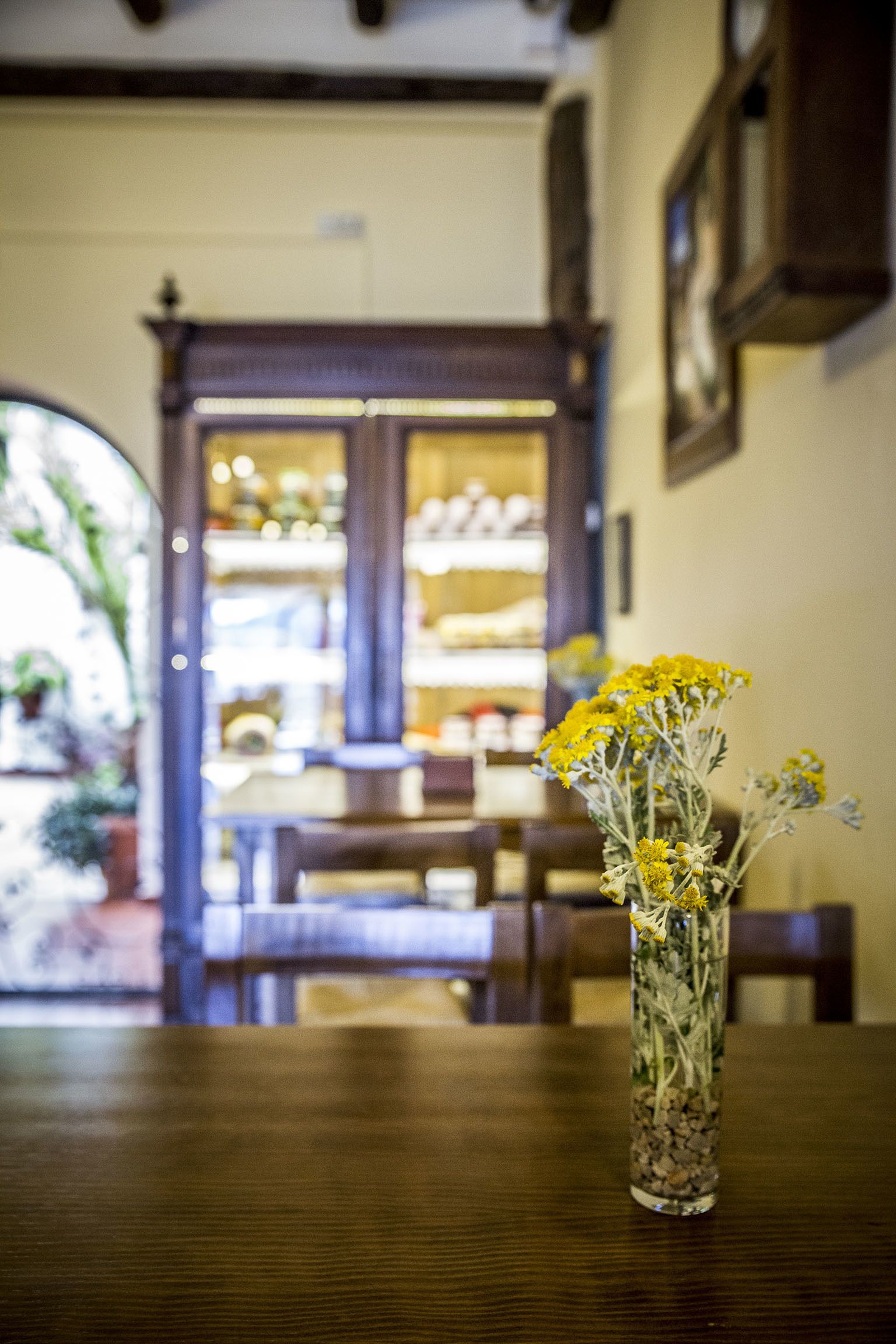 A wooden table with a vase of yellow flowers. In the background, there is a wooden cabinet with glass doors and a framed picture on the wall.