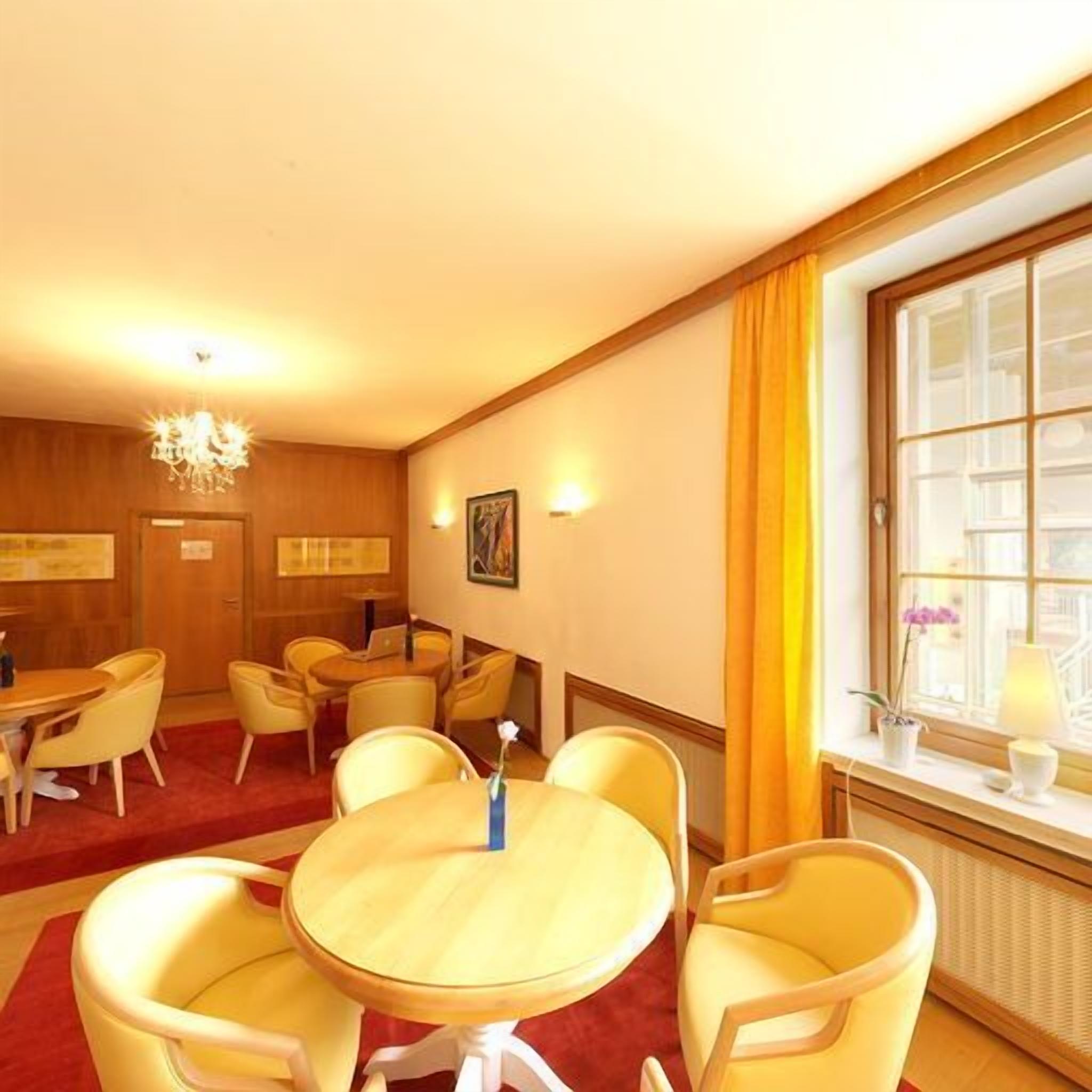 A hotel room with a dining area featuring round tables and yellow chairs, a chandelier, and a window with curtains.